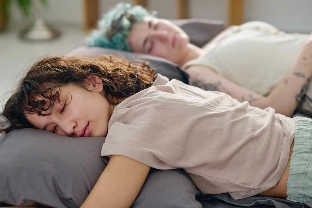 Young tired brunette woman in cotton t-shirt lying on soft grey pillow on double bed during deep sleep on background of her girlfriend
